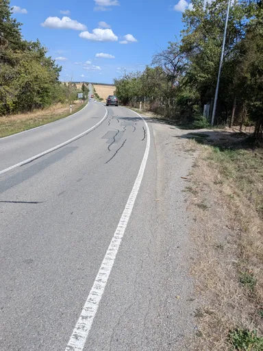 Silnice stoupající mezi poli a stromy, v dálce projíždějící auto