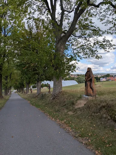 Asfaltová cesta vede alejí do města, vpravo u kmene stojí dřevěná socha, na obzoru střechy Bystřice nad Pernštejnem.