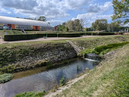 Vyzděné koryto potoka s malým stupněm a tůní, v pozadí hala zimního stadionu.