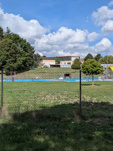 Modré bazény koupaliště za plotem, kolem travnatá plocha a herní prvky.
