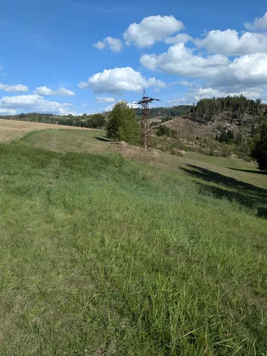 Travnatá cesta vede podél okraje pole, v pozadí sloup vysokého napětí a zalesněné kopce.