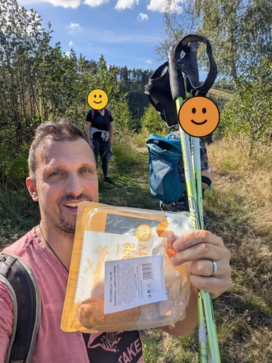 Selfie na lesní pěšině: muž drží balíček špekáčků, za ním stojí dva turisté s batohy a holemi, jejich obličeje jsou zakryté smajlíkem.