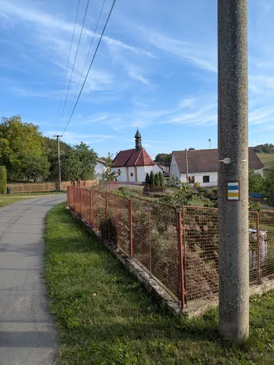 Chodník vede do vsi kolem plotu, v pozadí kaple s věžičkou a domy.