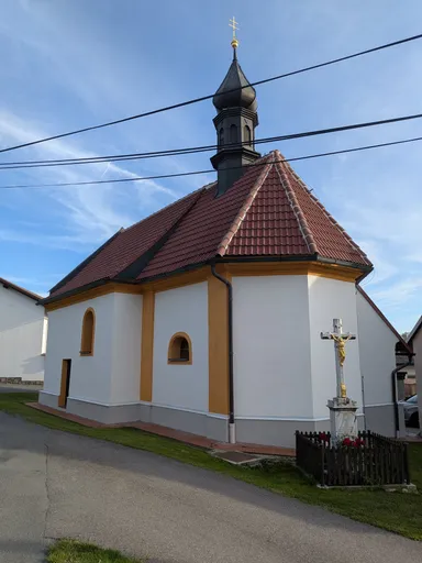 Bílá kaple s červenou střechou a černou věžičkou, vedle ní kamenný kříž.