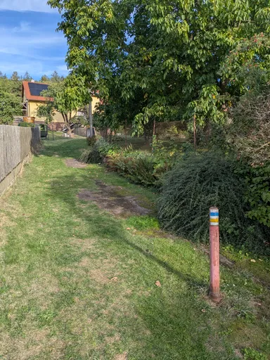 Travnatá cesta mezi ploty stoupá vsí, vpravo sloupek s turistickou značkou.