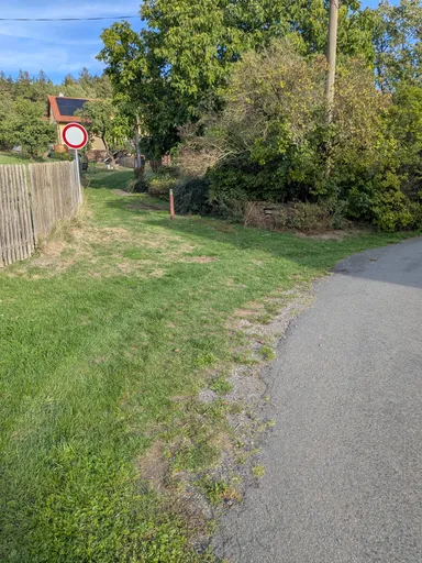Travnatý pás mezi plotem a asfaltovou cestou, na kraji značka zákazu vjezdu.