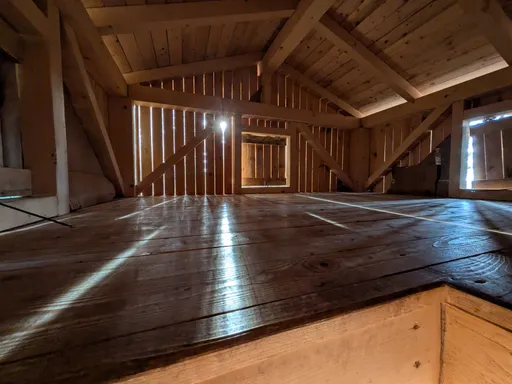 Vnitřek dřevěného přístřešku s prkennou podlahou, lavicemi a krovem.