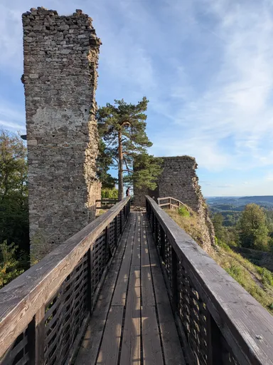 Dřevěná lávka se zábradlím vede podél vysoké kamenné zdi, vpravo otevřený výhled.