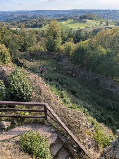 Pohled ze zříceniny do údolí s loukami a lesy, v popředí dřevěné zábradlí.