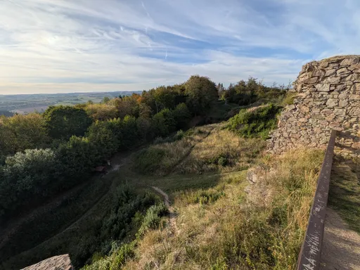 Hluboký příkop mezi zalesněným hřebenem a hradní zdí, v pozadí krajina.