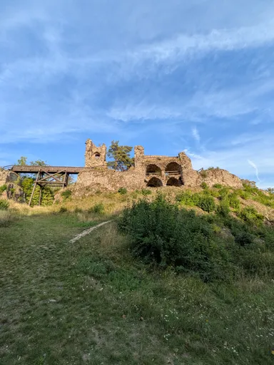 Zřícenina hradu s klenbami a dřevěnou lávkou na travnatém návrší pod modrou oblohou.