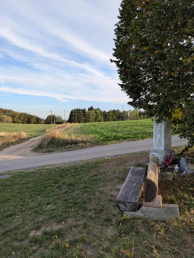 Kamenný pomníček s květinami a kamennou lavicí pod stromem u rozcestí.