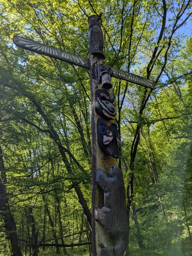 Vyřezávaný dřevěný totem s křížem a postavami uprostřed jarního lesa.
