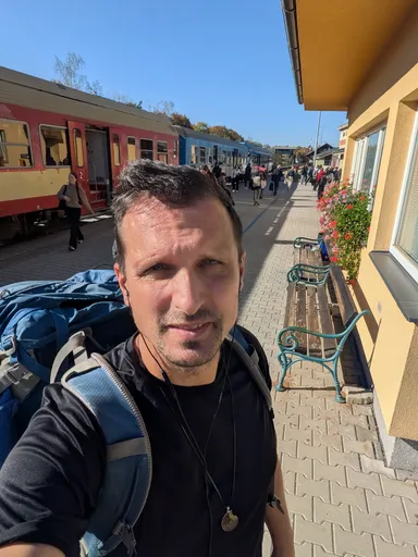 Poutník s batohem si dělá selfie na nástupišti nádraží v Náměšti nad Oslavou, za ním stojí vlak.