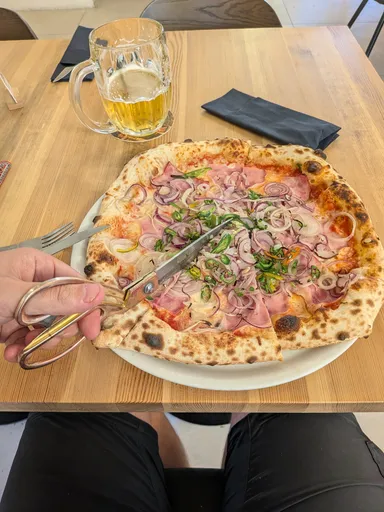 Neapolská pizza se šunkou a cibulí na stole, vedle leží nůžky na krájení a stojí sklenice piva.