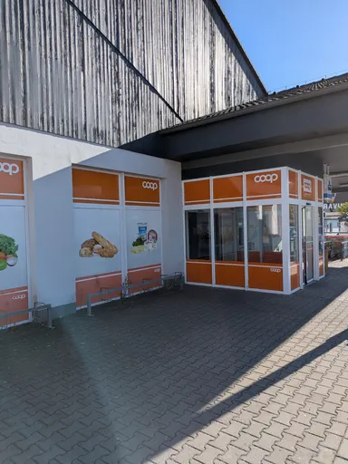 Vchod do prodejny COOP s oranžovými reklamními panely pod dřevěnou fasádou.
