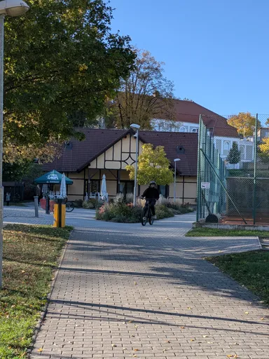Dlážděná cesta k hrázděnému občerstvení u sportovišť, po ní projíždí cyklista.