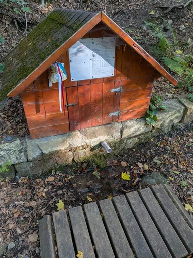 Malá dřevěná stříška studánky s cedulí a českou vlajkou, před ní dřevěný rošt.
