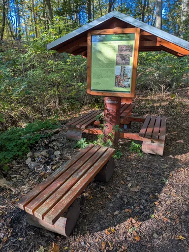 Dřevěný přístřešek s informační tabulí a lavicemi v lese u studánky.