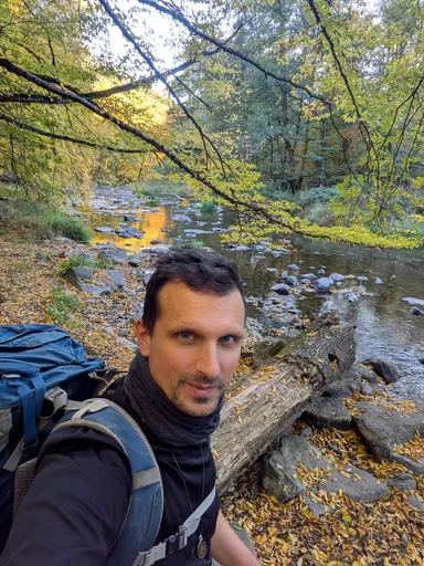 Poutník s batohem si dělá selfie u kamenitého koryta řeky Oslavy v podzimním lese.