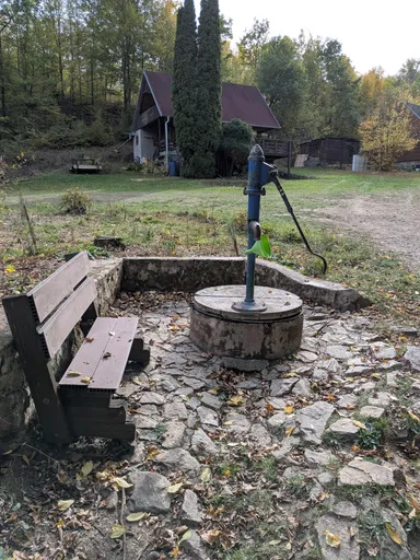 Studna s ruční pumpou a lavičkou na dlážděném plácku u chaty.